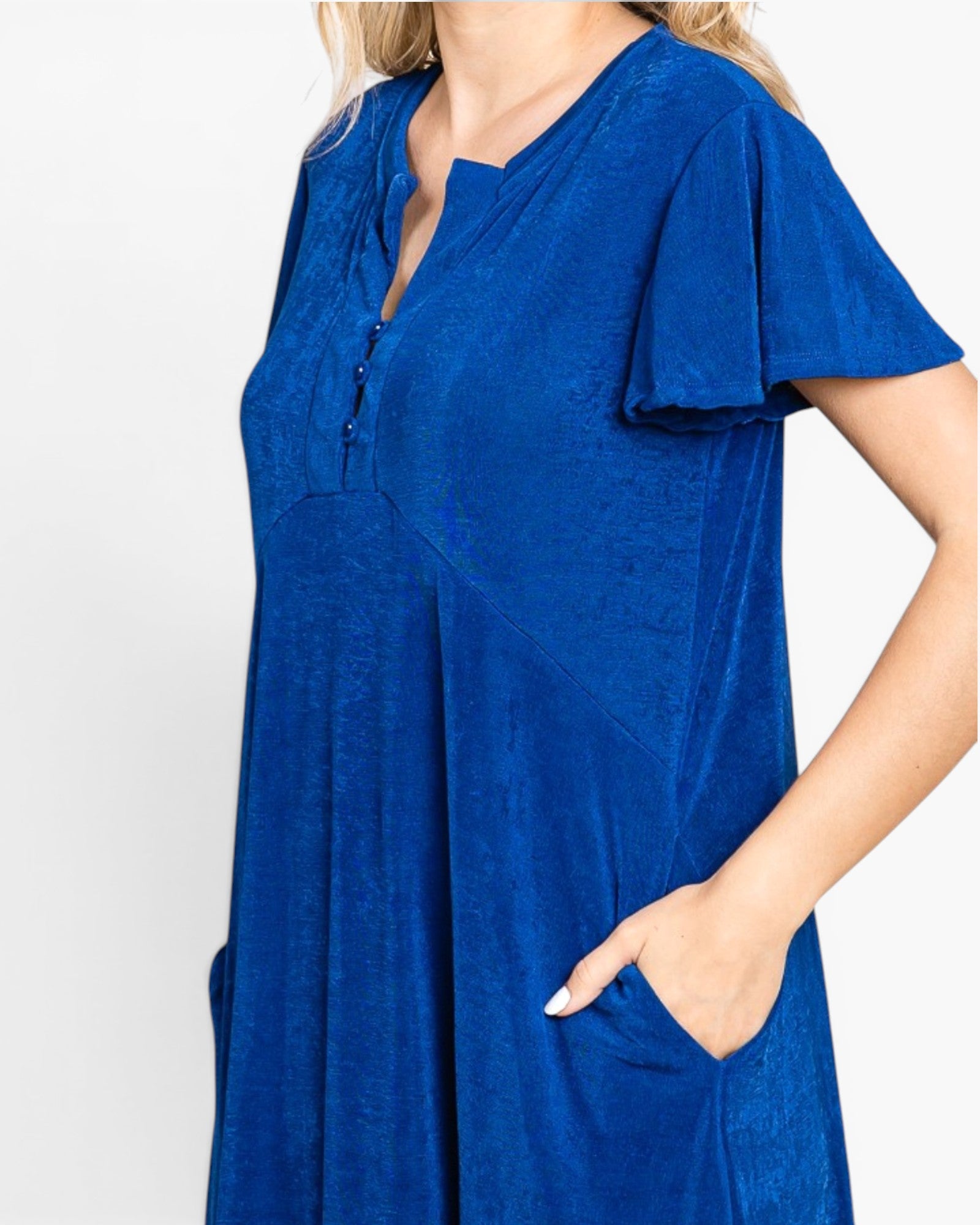 A woman wearing a royal blue short-sleeve tunic dress with a split V-neckline and side pockets, shown against a plain light gray background.