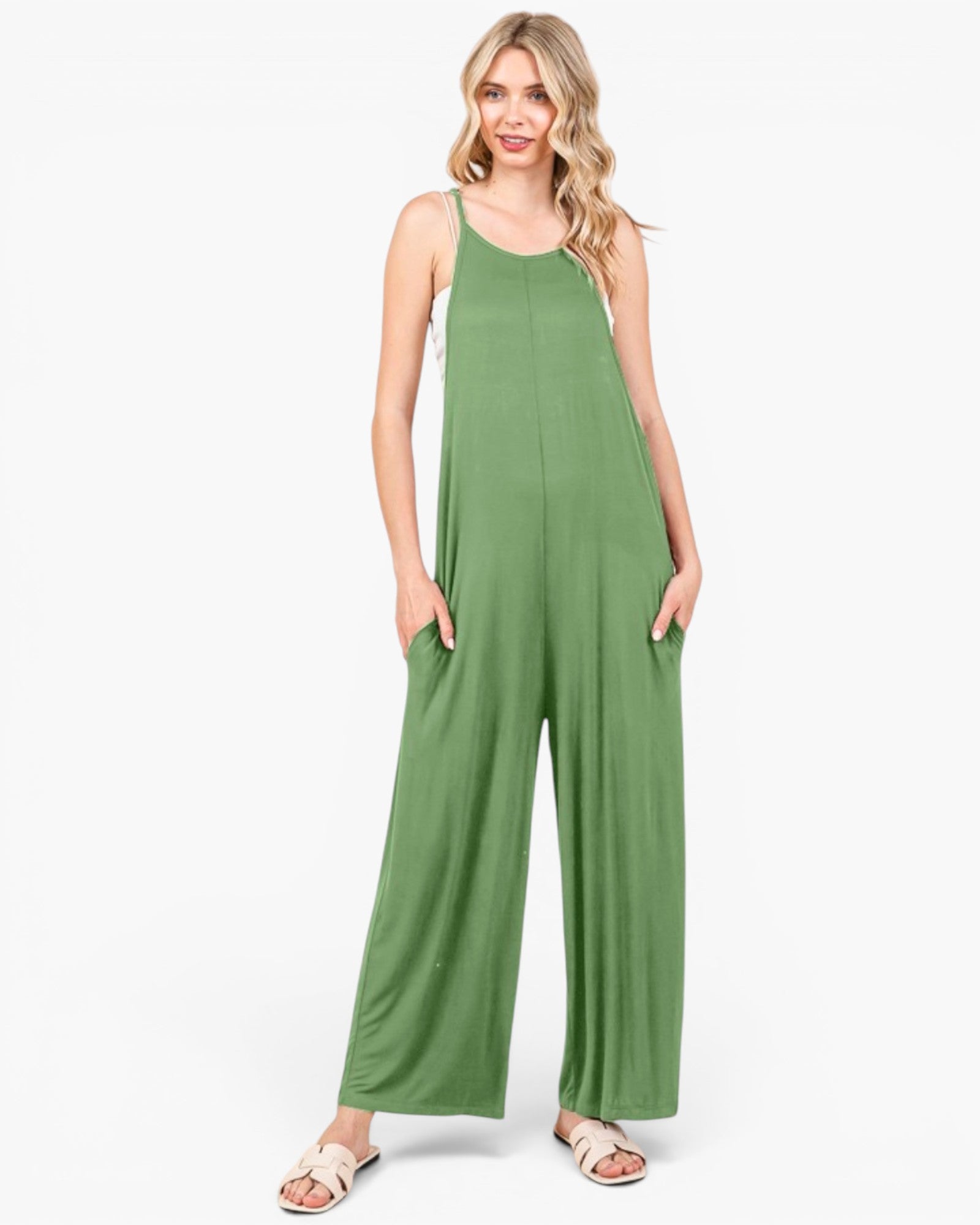 A woman modeling a sage green sleeveless jumpsuit with wide legs and side pockets, paired with light pink sandals, against a plain white studio background.