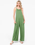 A woman modeling a sage green sleeveless jumpsuit with wide legs and side pockets, paired with light pink sandals, against a plain white studio background.