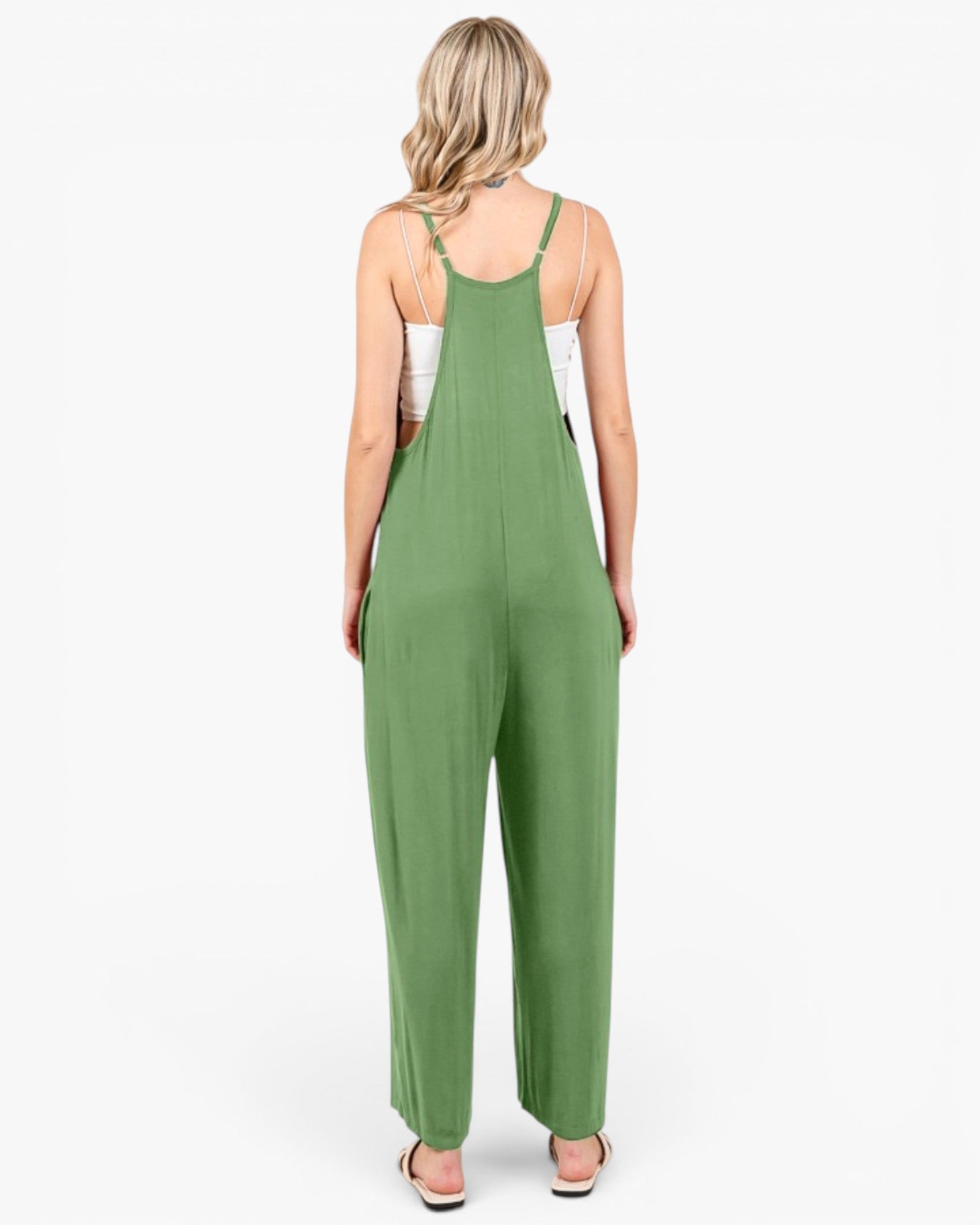 A woman viewed from behind wearing a sage green sleeveless jumpsuit with wide legs and thin shoulder straps, paired with a white tank top and white slide sandals, against a plain white background.