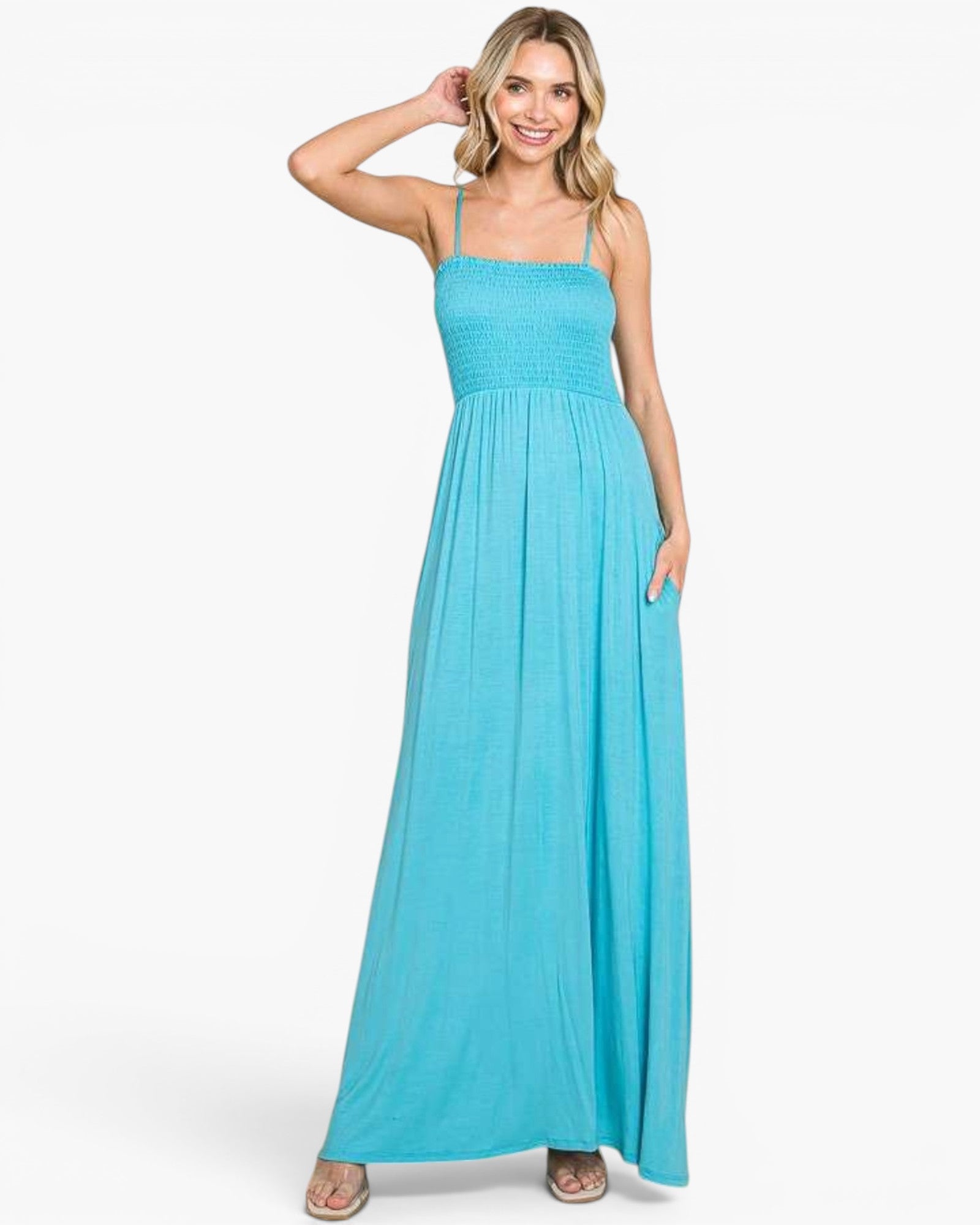 A woman modeling a bright turquoise blue maxi dress with thin spaghetti straps, a smocked bodice, and a flowing skirt, set against a plain white studio background.