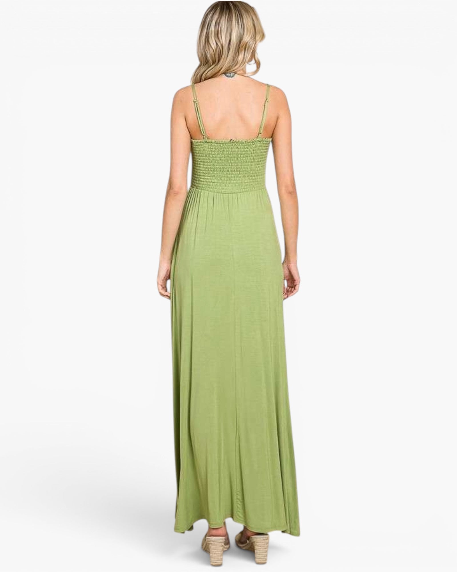 A woman viewed from the back wearing a sage green maxi dress with thin spaghetti straps, a smocked bodice, and a flowing skirt, paired with beige high-heeled sandals against a light gray studio background.
