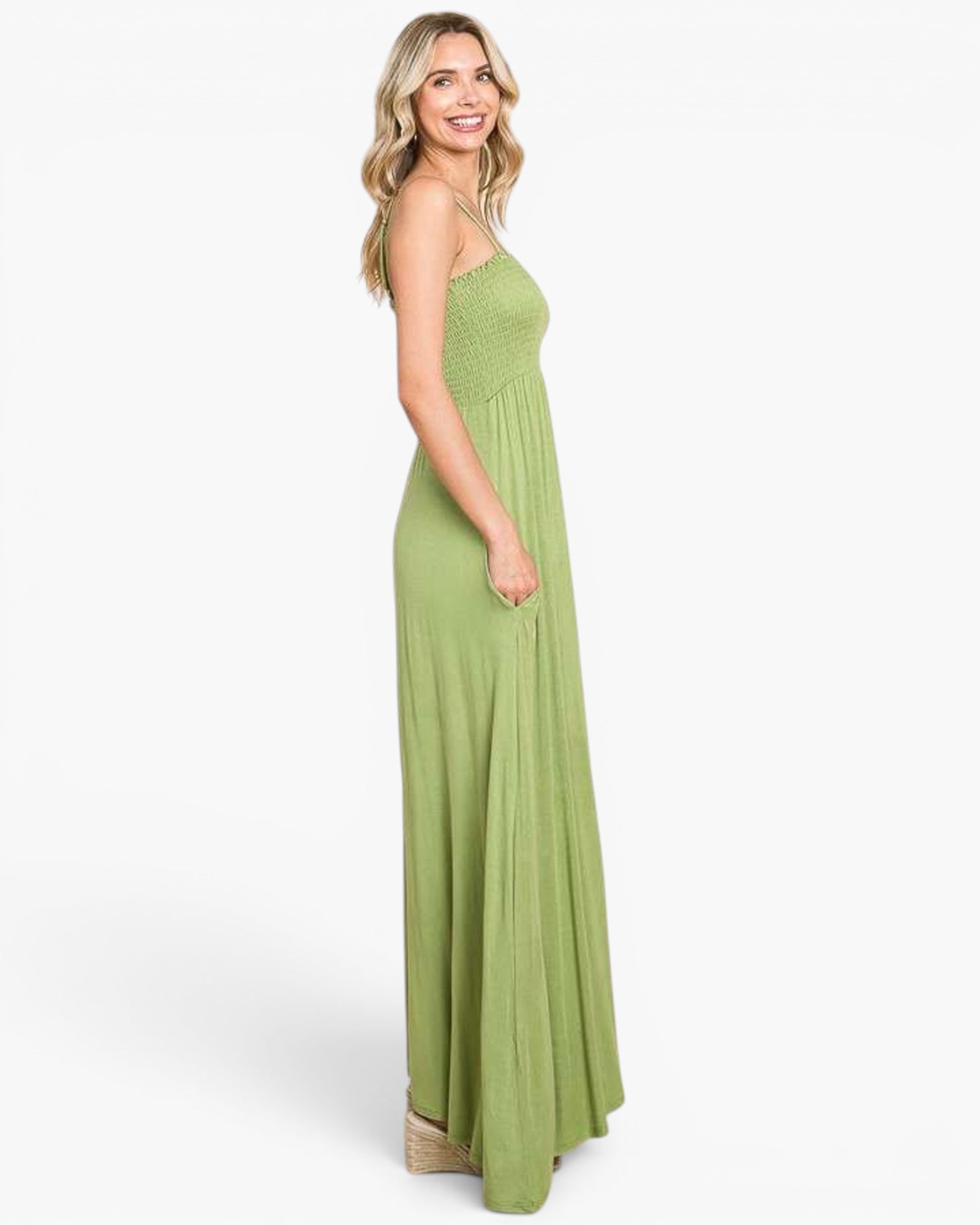 A woman modeling a sleeveless, smocked bodice, floor-length sage green maxi dress against a plain white studio background.