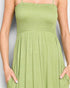 A close-up of a woman wearing a light green smocked spaghetti-strap dress, showing the smocked bodice, gathered waist, and pleated skirt with side pockets.