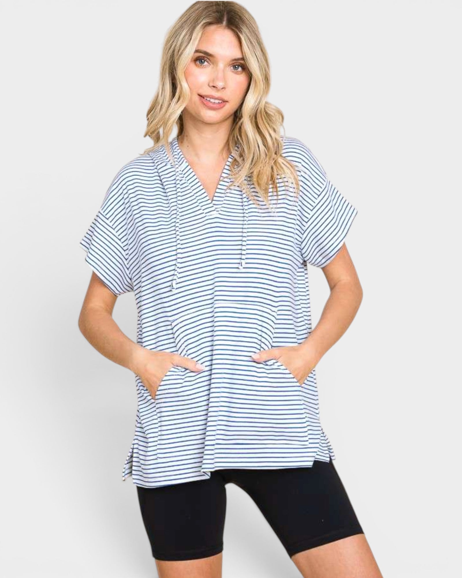 A woman modeling a short-sleeve striped tunic top with drawstrings and front pockets, paired with black bike shorts, against a light gray studio background.