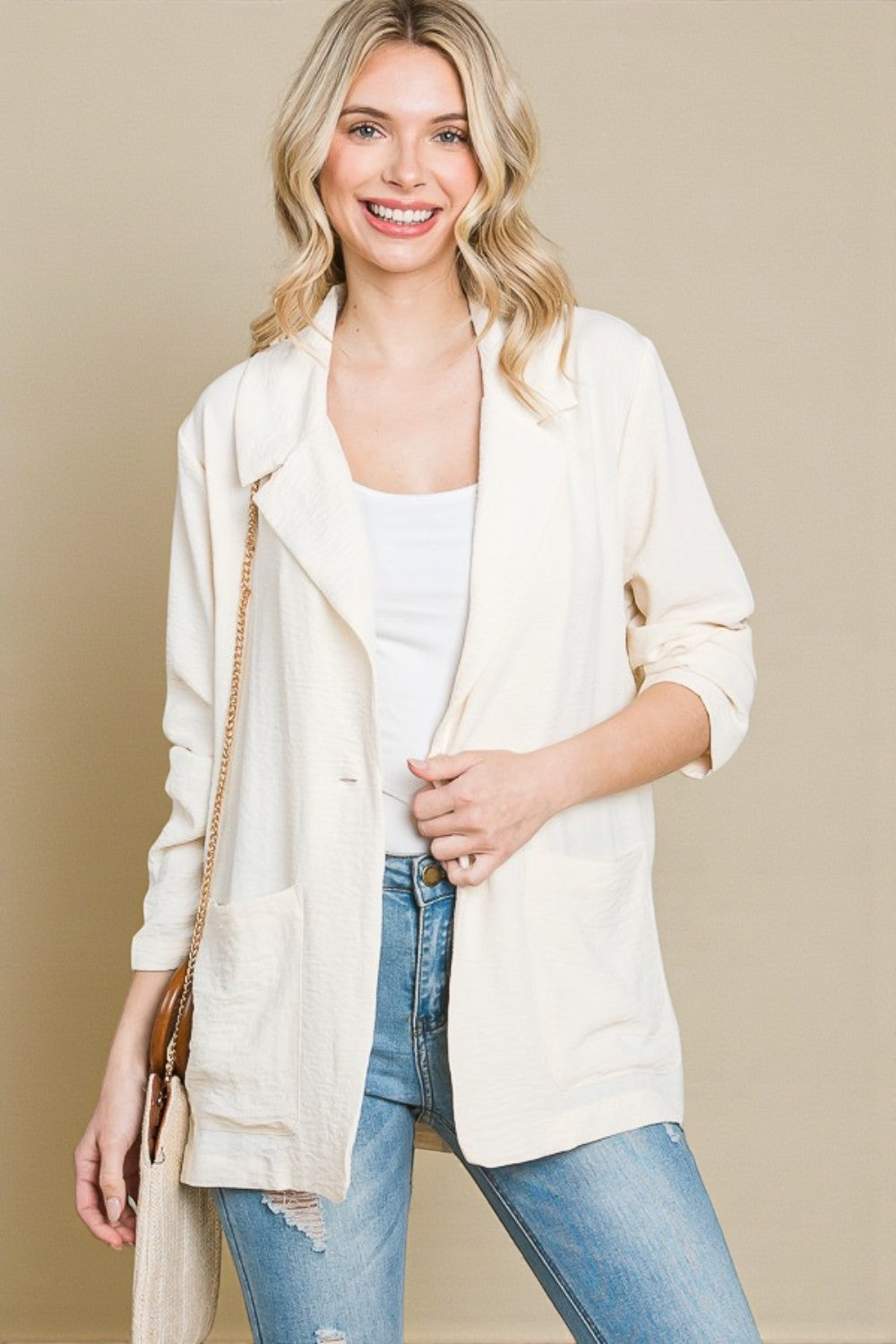 Woman wearing a cream cardigan over a white top and blue jeans, standing against a beige background.