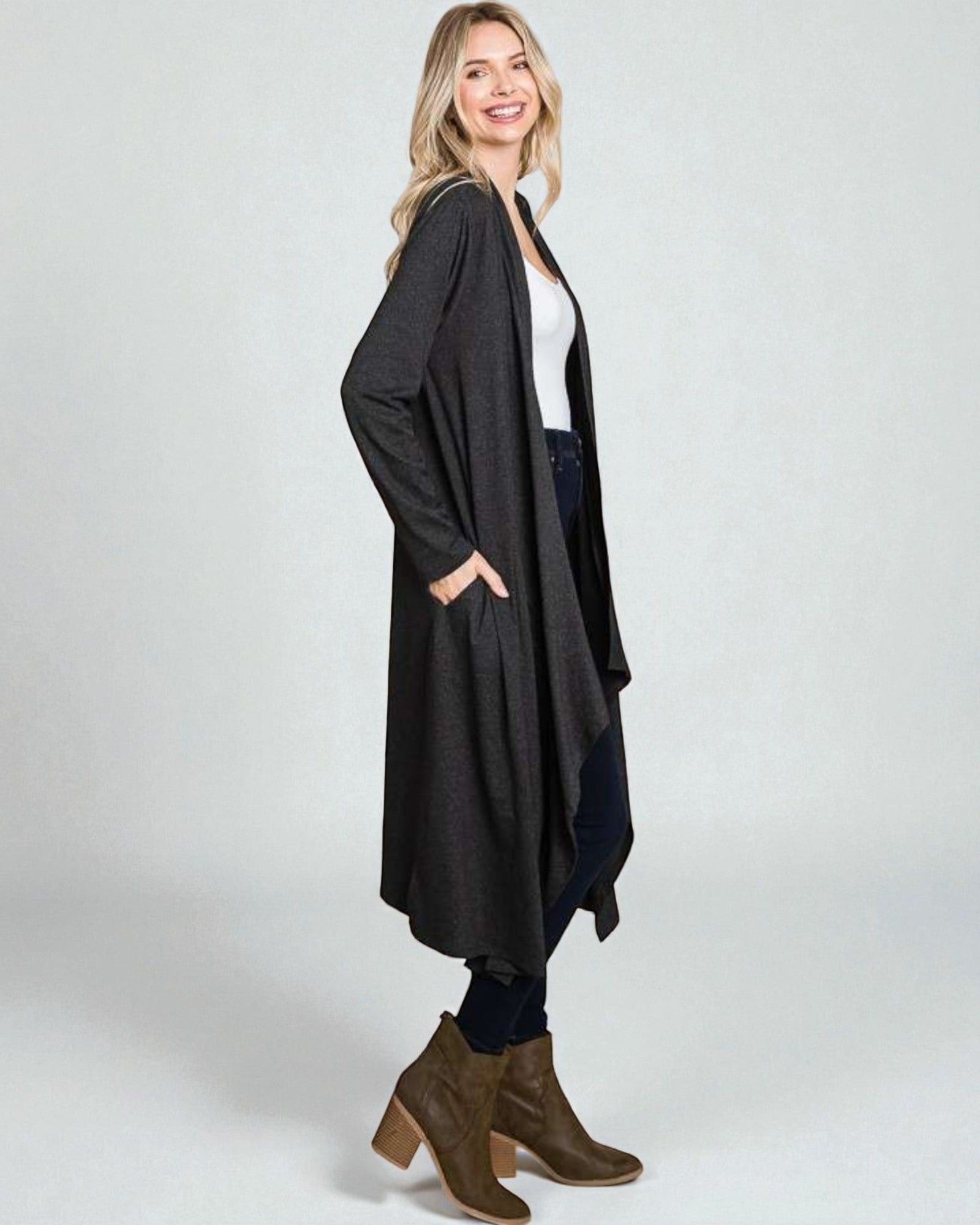 Woman wearing a long dark gray cardigan over a white top and jeans, standing against a plain background.