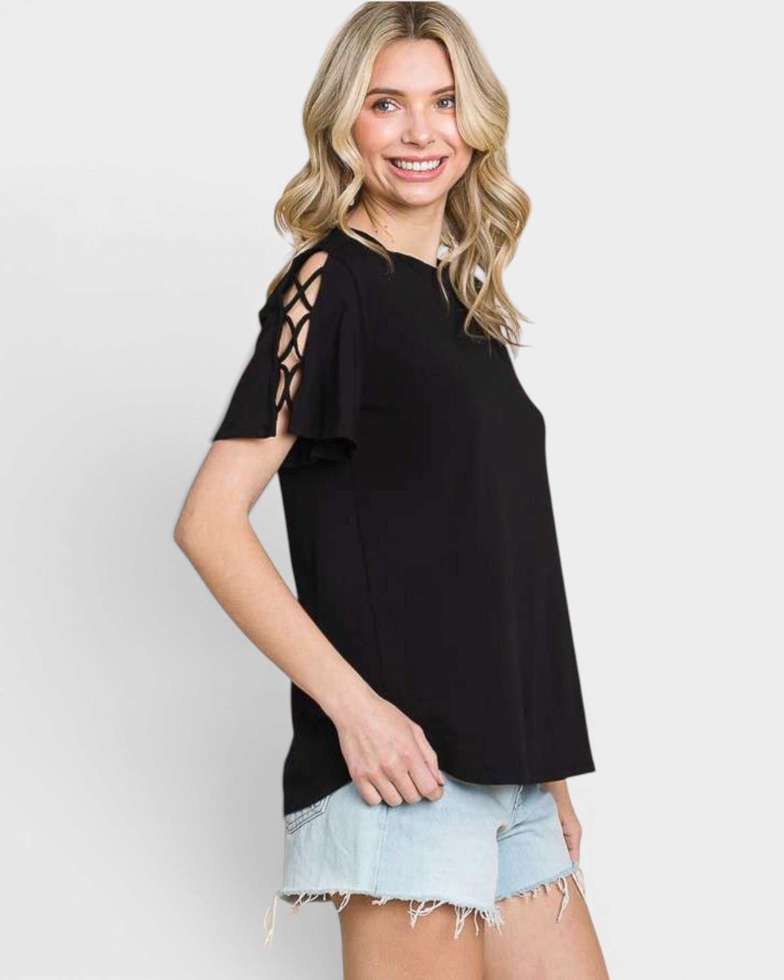 A woman wearing a black short-sleeve top with lace-up cutout details on the shoulders, paired with light-wash denim shorts, against a light gray studio background.