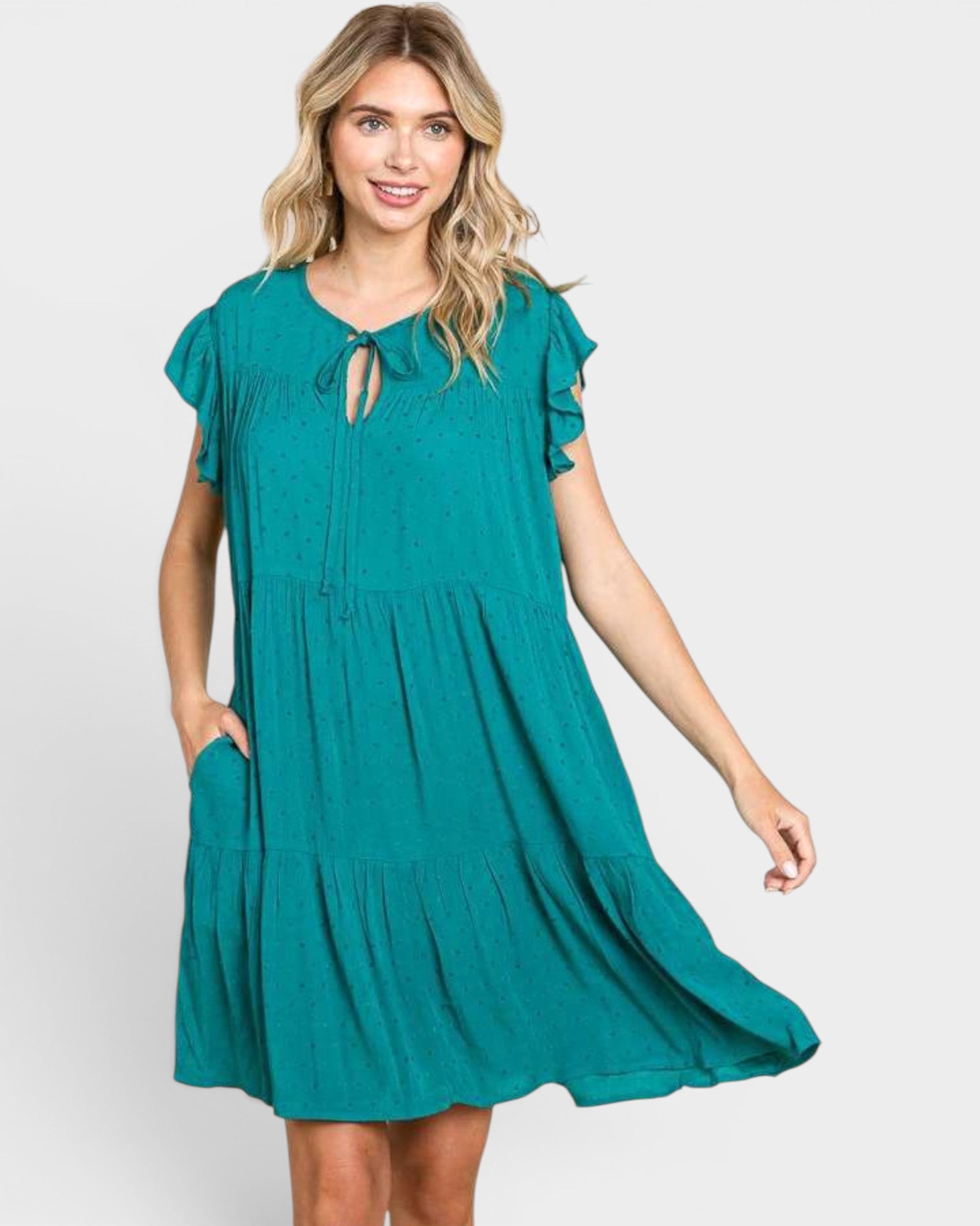 A woman modeling a teal green tiered mini dress with ruffled short sleeves, a keyhole tie neckline, and side pockets, set against a plain light gray background.