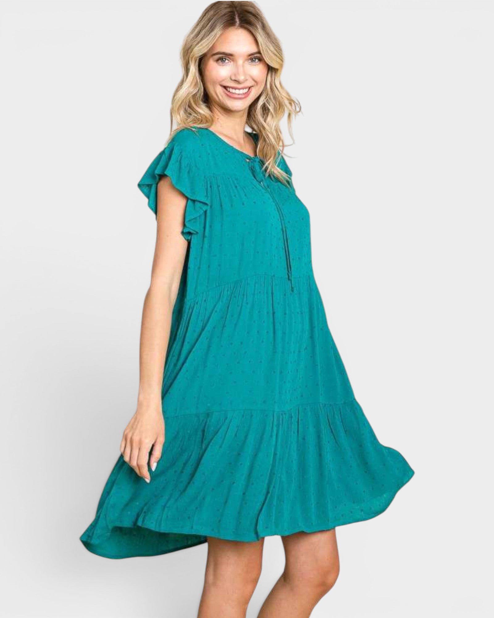 A woman modeling a teal green tiered mini dress with ruffled short sleeves and a gathered waist, photographed against a light gray studio background.