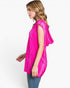 A woman shown in side profile wearing a bright hot pink sleeveless tunic top with draped, wide shoulders and blue jeans, against a plain white studio background.