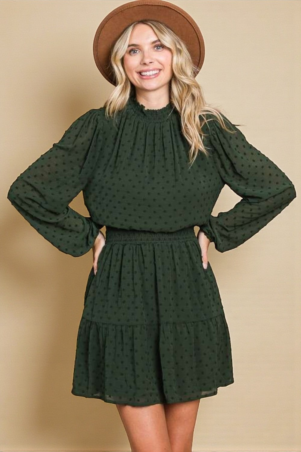 Woman wearing a green dress with long sleeves and a brown hat against a beige background