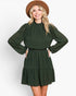 A woman modeling a dark green long-sleeve mini dress with a subtle polka dot pattern, paired with a brown wide-brim hat, against a plain light gray background.