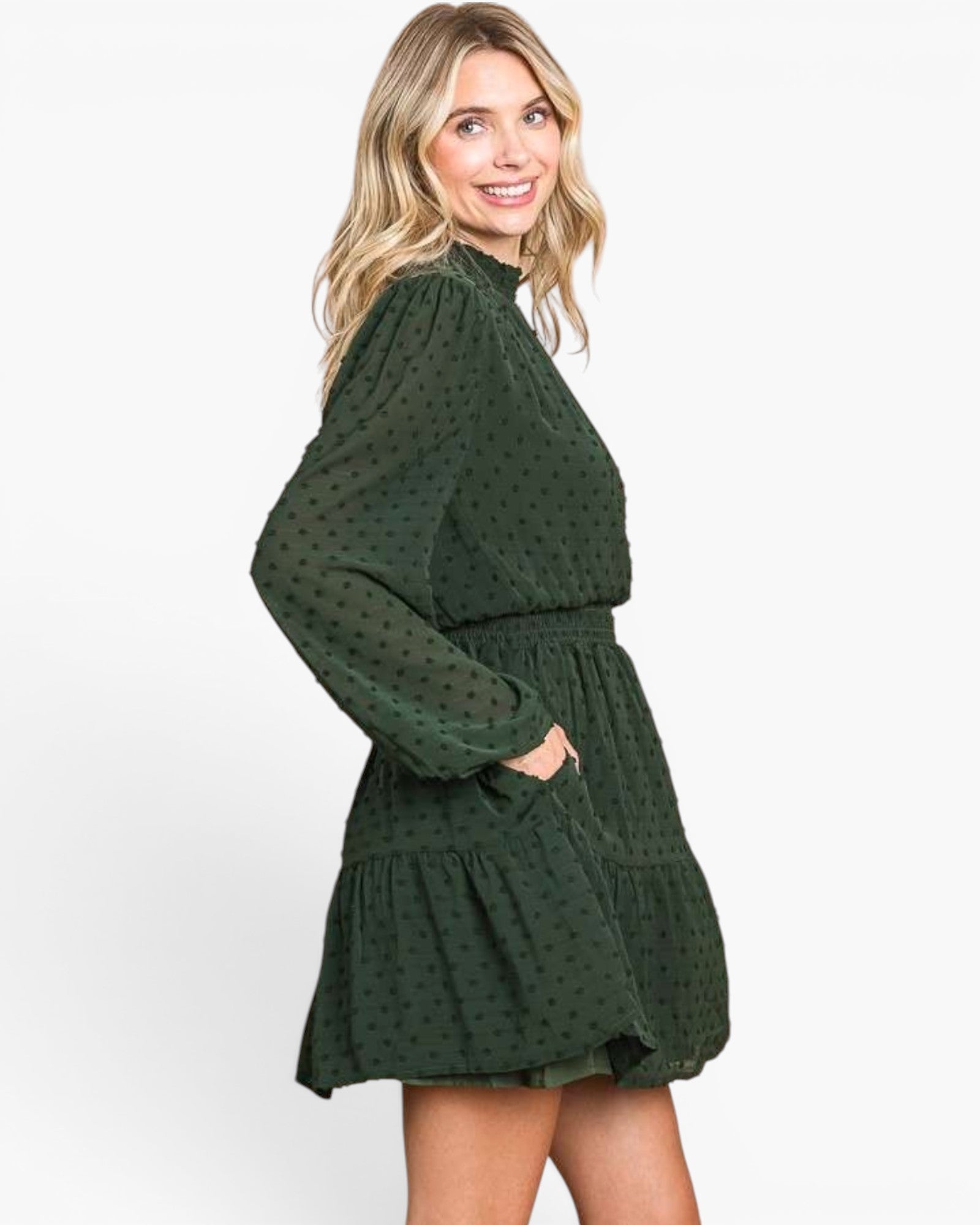 A woman modeling a dark green long-sleeve mini dress with a subtle polka dot pattern, high mock neck, and a tiered skirt, set against a plain white studio background.