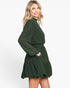 A woman modeling a dark green long-sleeve mini dress with a subtle polka dot pattern, high mock neck, and a tiered skirt, set against a plain white studio background.