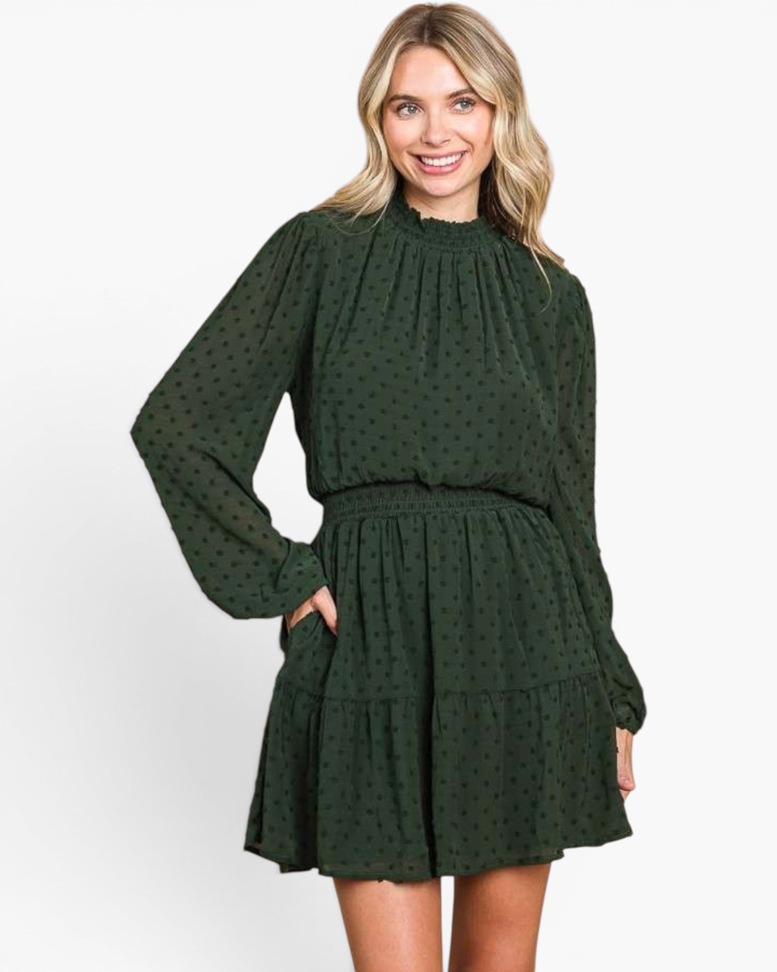 A woman modeling a dark green long-sleeve mini dress with a subtle polka dot pattern, high gathered neckline, and tiered skirt against a plain white background.