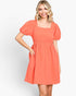 A woman modeling a short puff-sleeve dress in a warm coral orange color with a square neckline and gathered waist, set against a plain light gray studio background.