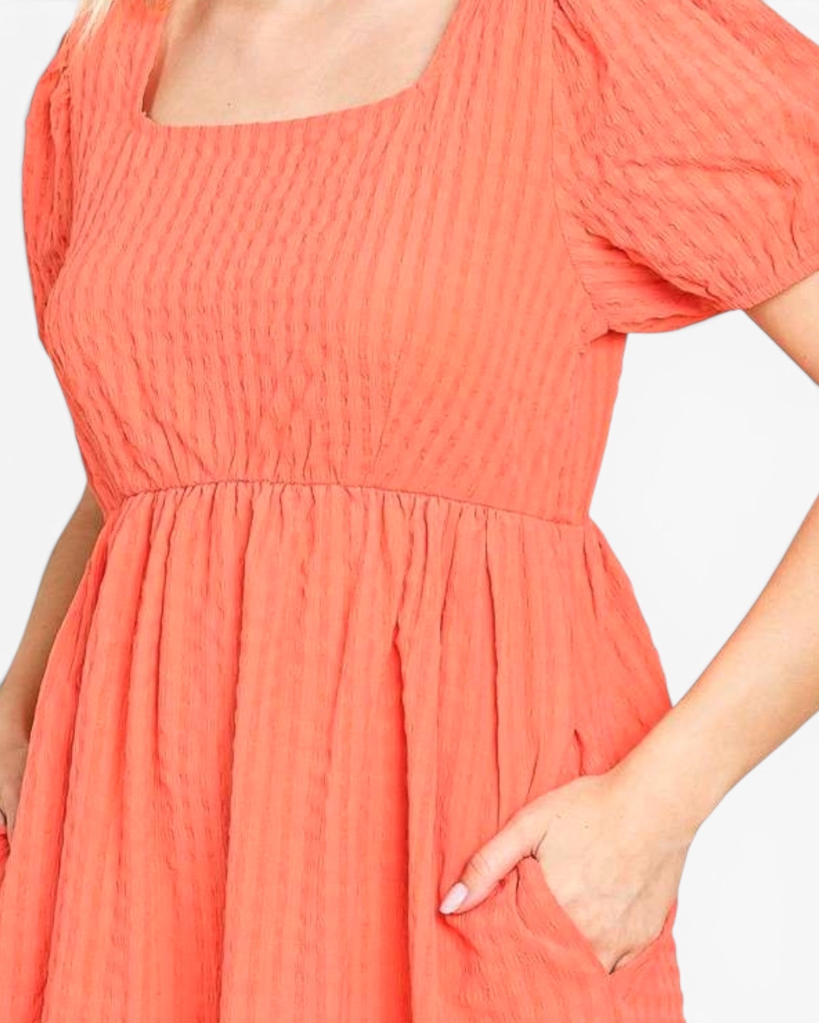 A close-up of a woman wearing a coral orange textured dress with a square neckline, short puff sleeves, and a gathered skirt, shown against a plain light background.