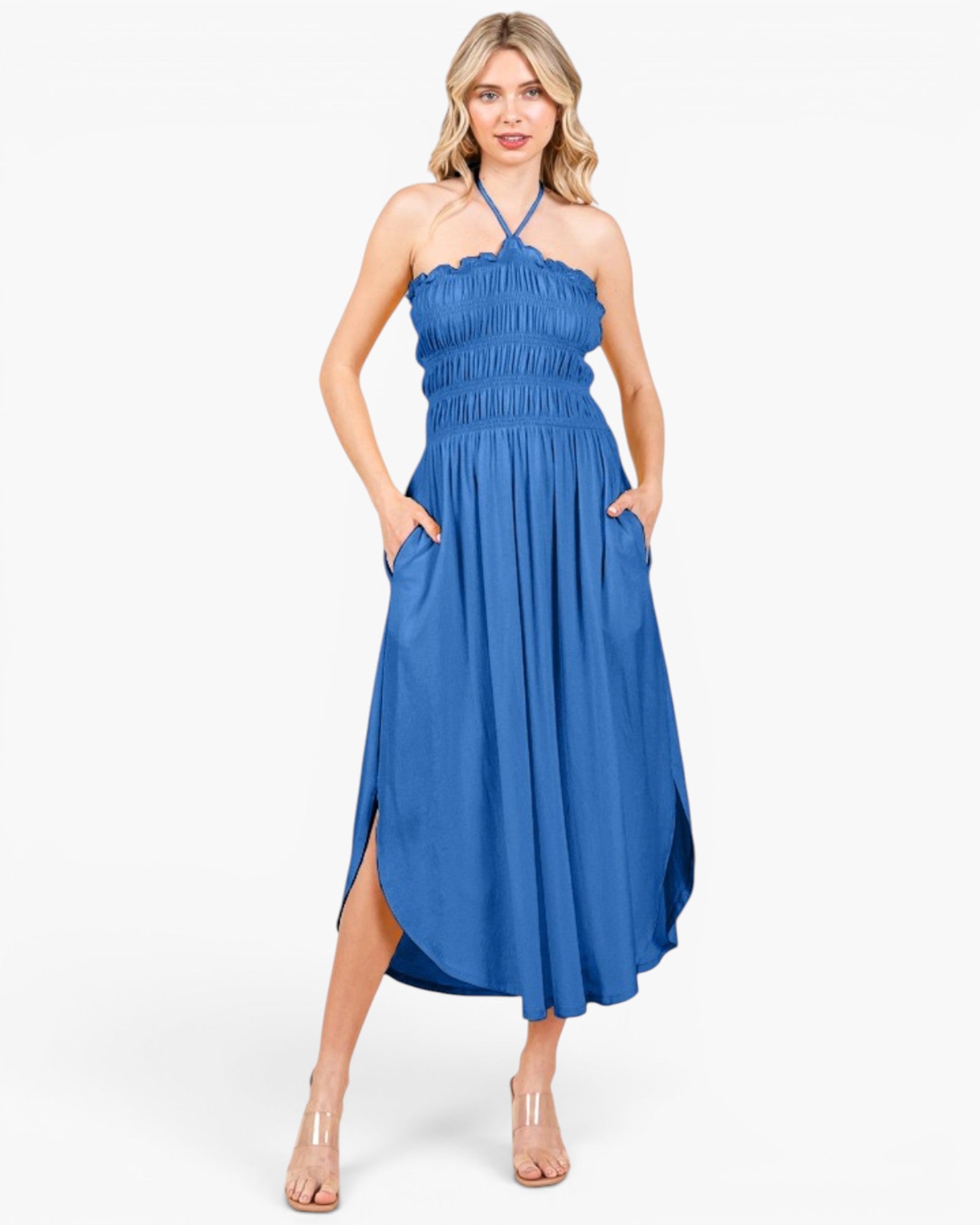 A woman modeling a blue halter-neck maxi dress with a smocked bodice, side pockets, and a high-low skirt, paired with clear high-heeled sandals against a light gray studio background.