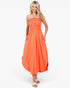 A woman modeling a bright orange halter-neck maxi dress with a smocked bodice and side pockets, paired with light pink sandals, against a plain white studio background.