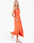 A woman modeling a bright orange halter-neck midi dress with a smocked bodice, side pockets, and a flowing skirt, paired with beige slide sandals against a plain white studio background.