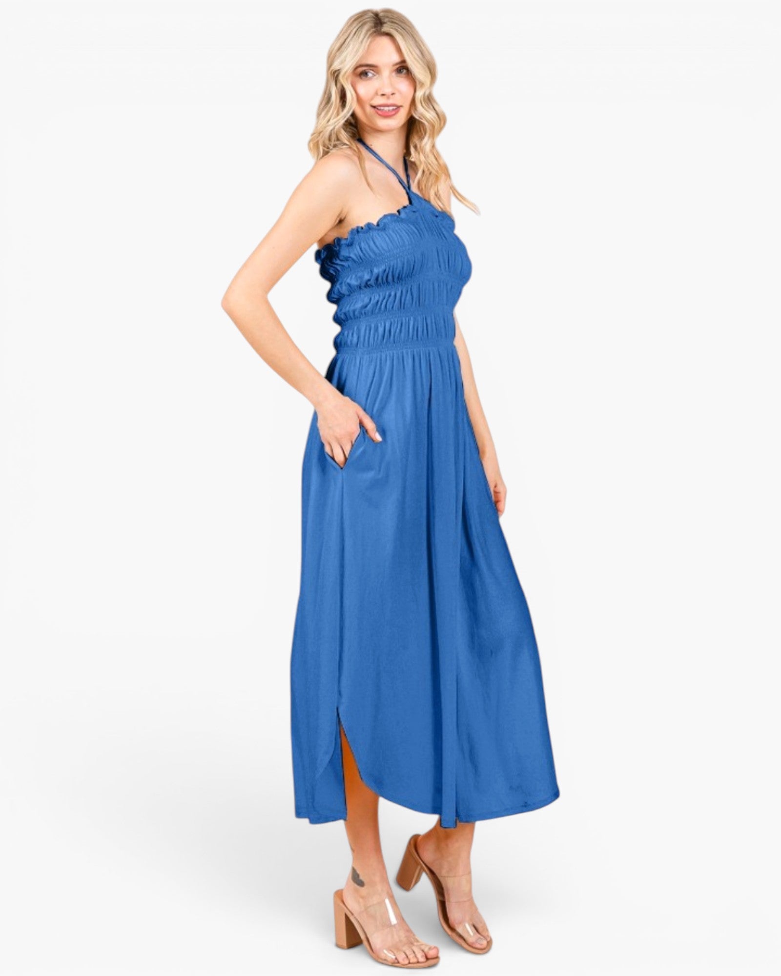 A woman modeling a blue halter-neck midi dress with a gathered waist and side pockets, paired with beige heeled sandals, against a plain white studio background.
