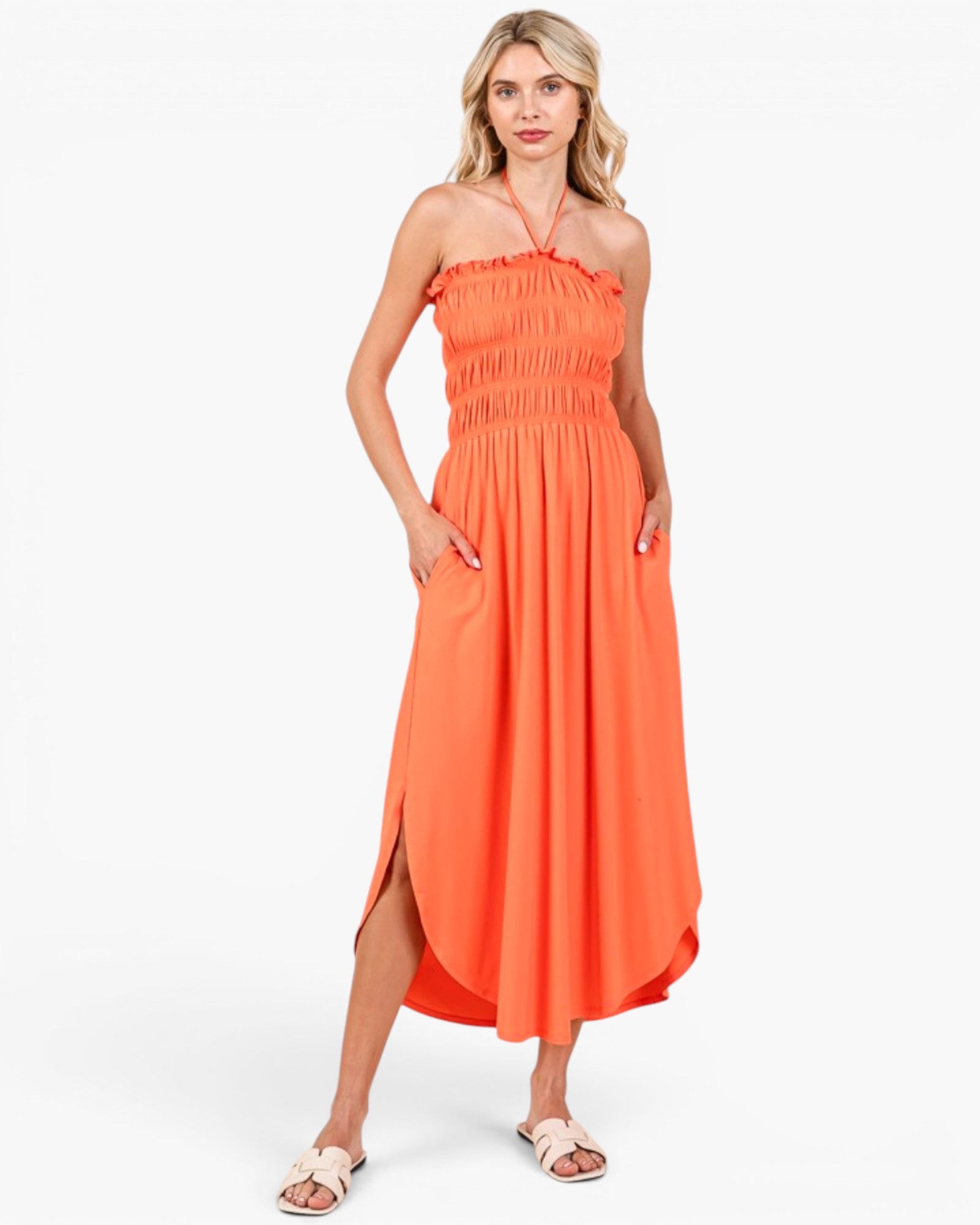A woman modeling a bright orange halter-neck midi dress with a smocked bodice and side slit, paired with light pink slide sandals, against a plain white studio background.