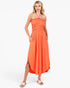 A woman modeling a bright orange halter-neck midi dress with a smocked bodice and side slit, paired with light pink slide sandals, against a plain white studio background.