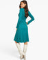 A woman modeling a teal long-sleeve midi dress with a flared skirt, paired with off-white knee-high boots, shown from a three-quarters back angle against a plain white studio background.
