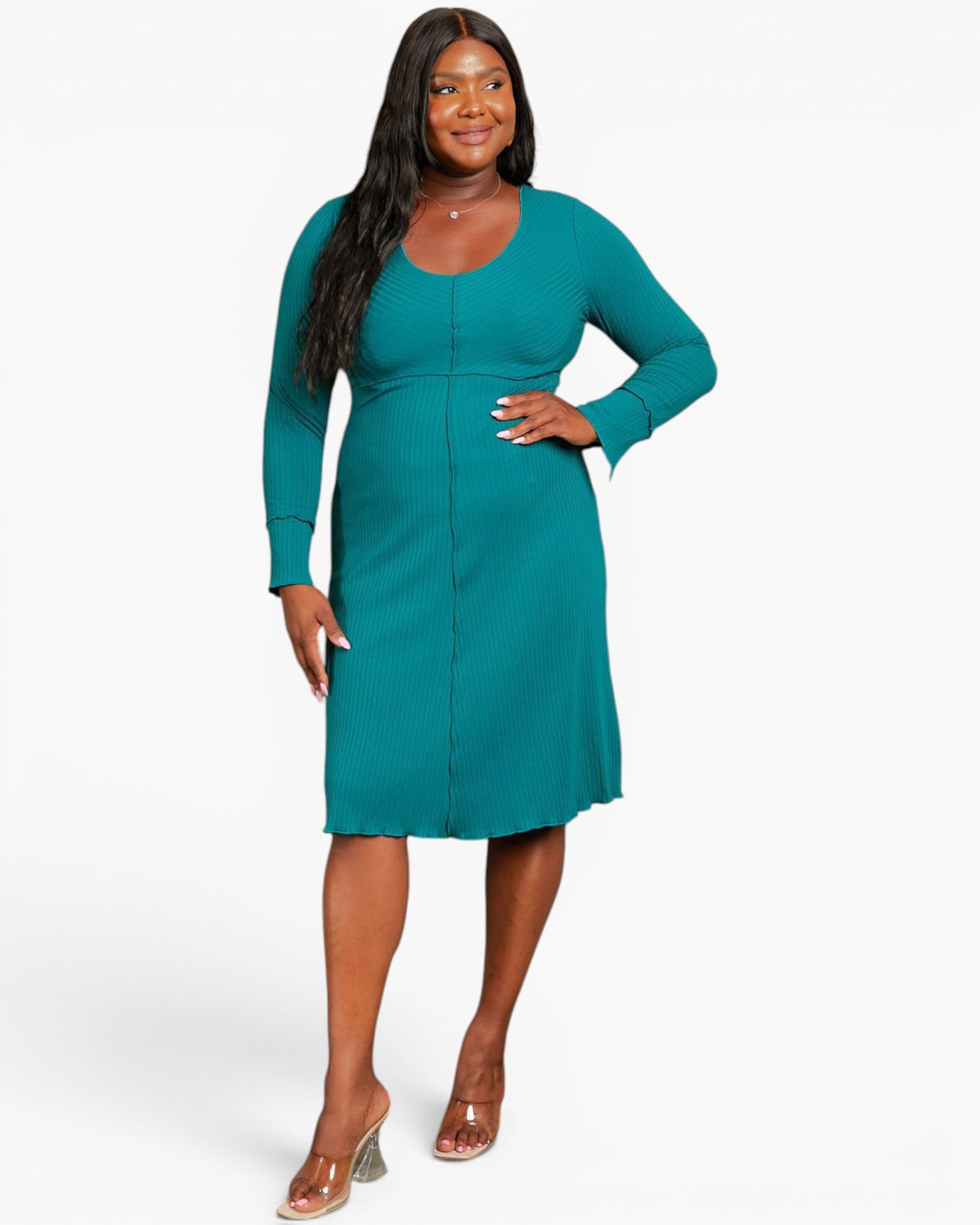 A woman modeling a teal long-sleeve knit dress with a scoop neckline and front button details, paired with clear high-heeled sandals, against a light gray studio background.