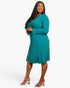 A woman modeling a teal long-sleeve ribbed knit midi dress with a V-neck and flared hem, paired with clear high-heel mules, against a plain light gray studio background.