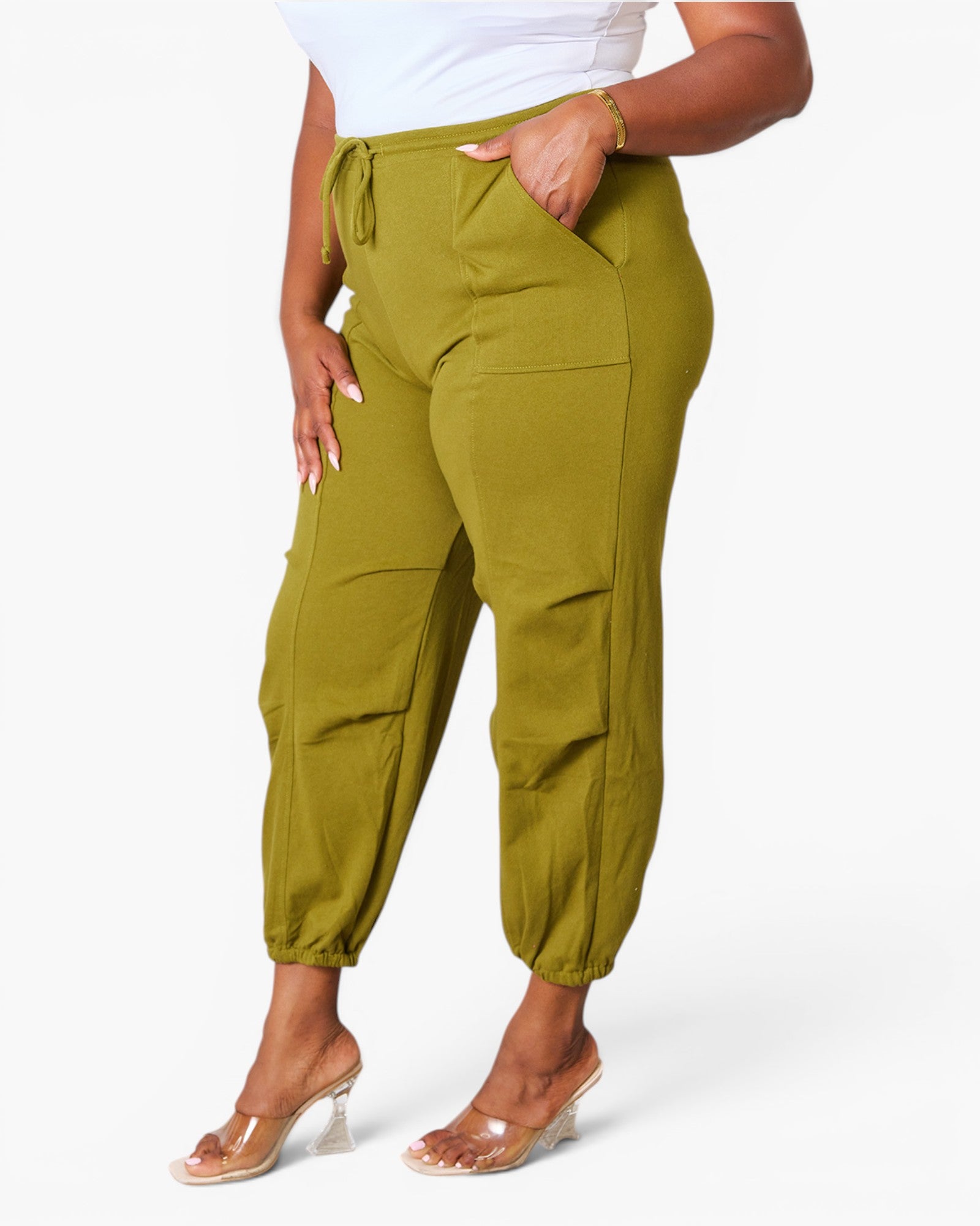 A model wearing olive green jogger-style pants with a drawstring waist, side pockets, and elastic cuffs, paired with a white top and clear high-heel sandals against a light gray studio background.