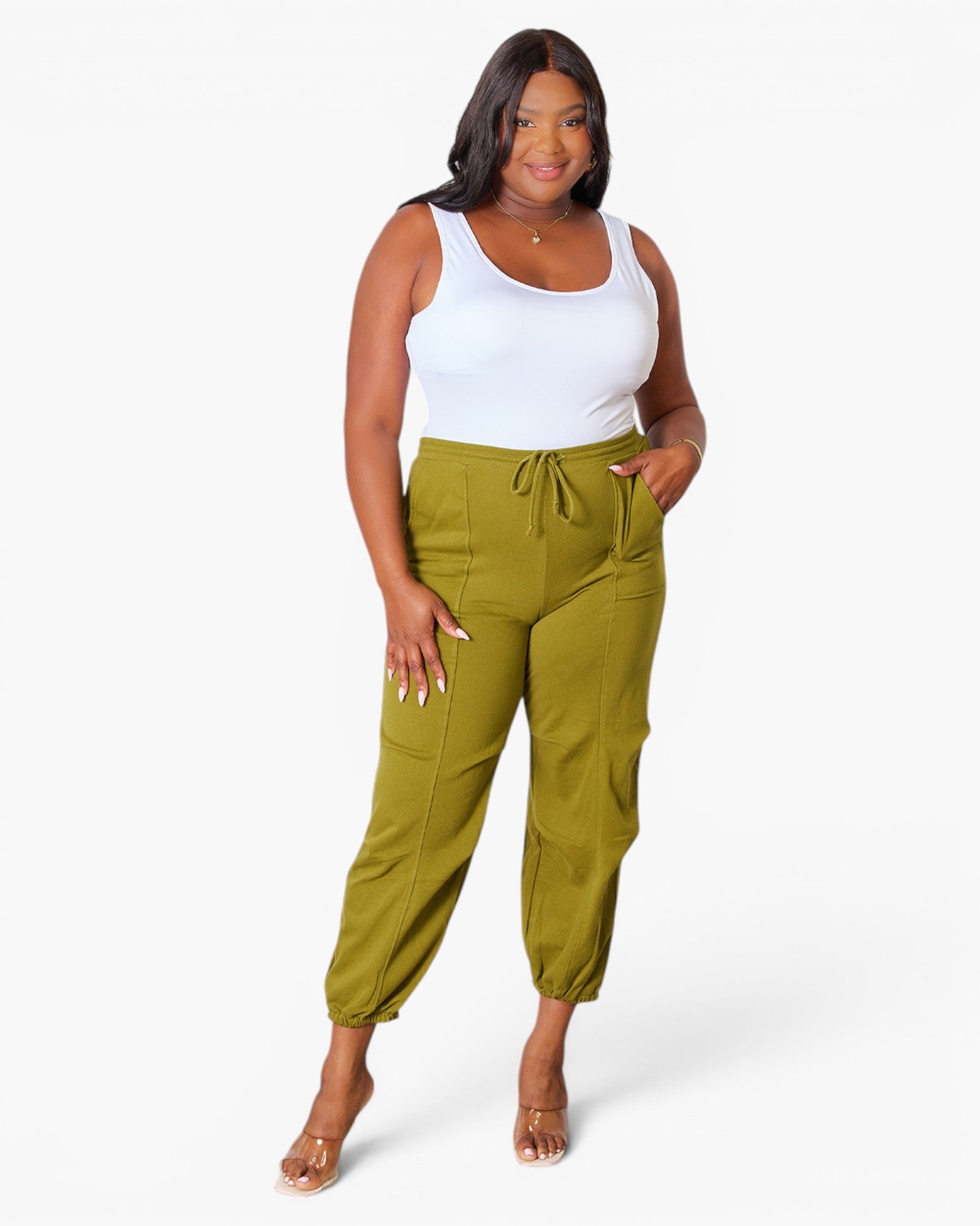 A woman modeling a pair of olive green jogger-style pants with a drawstring waist and side cargo pockets, paired with a white tank top, on a light gray studio background.