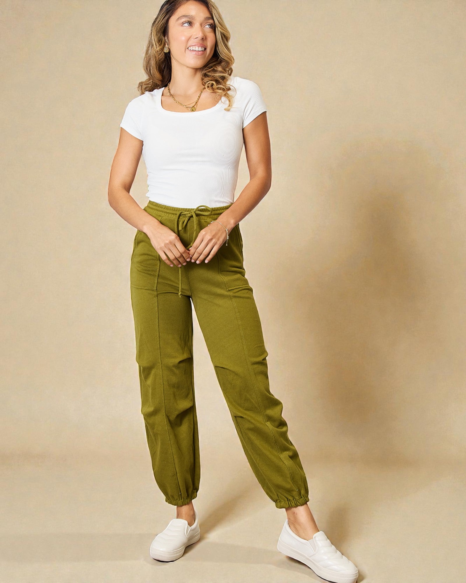 Woman wearing a white top and green pants against a beige background