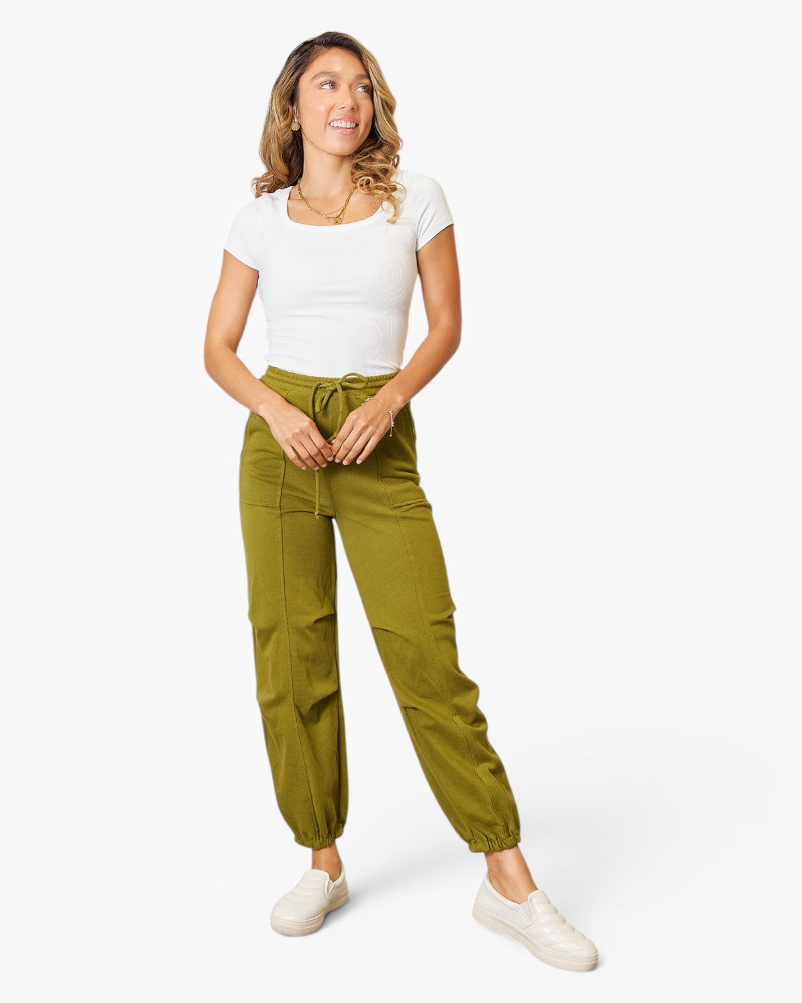 A woman modeling olive green jogger pants with a drawstring waist, paired with a white t-shirt and white sneakers, against a plain white studio background.