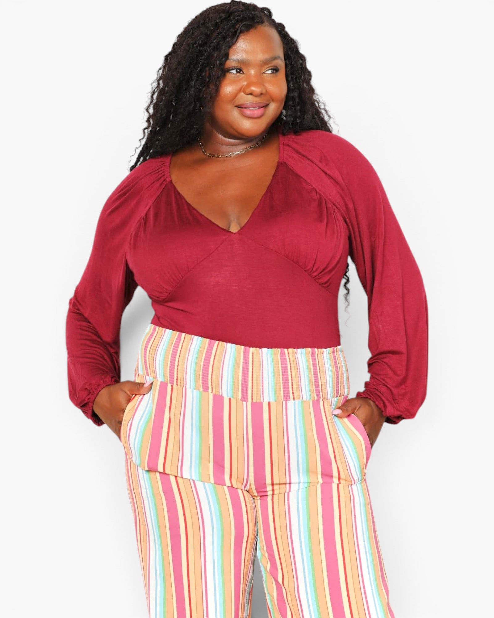 A woman modeling a long-sleeve wrap-style top in deep burgundy paired with pastel pink, yellow, and blue striped pants, against a plain light gray background.