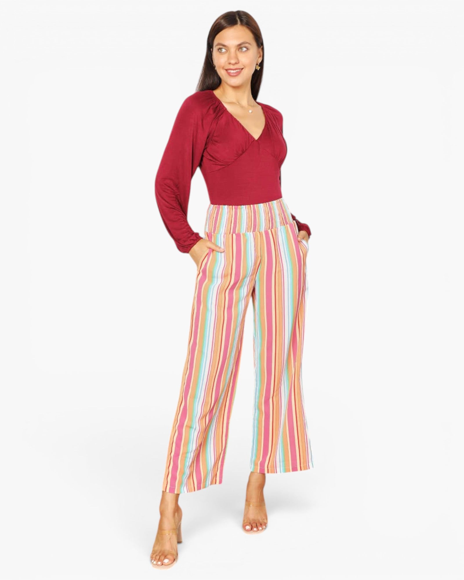 A woman modeling a long-sleeve wrap-style top in deep red paired with high-waisted cropped pants featuring vertical pastel stripes in pink, yellow, blue, and white, against a plain light gray studio background.
