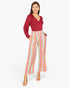 A woman modeling a long-sleeve wrap-style top in deep red paired with high-waisted cropped pants featuring vertical pastel stripes in pink, yellow, blue, and white, against a plain light gray studio background.