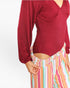 A close-up of a model wearing a burgundy long-sleeve bodysuit with a ruched chest and a cutout at the waist, paired with pastel striped pants against a plain white background.