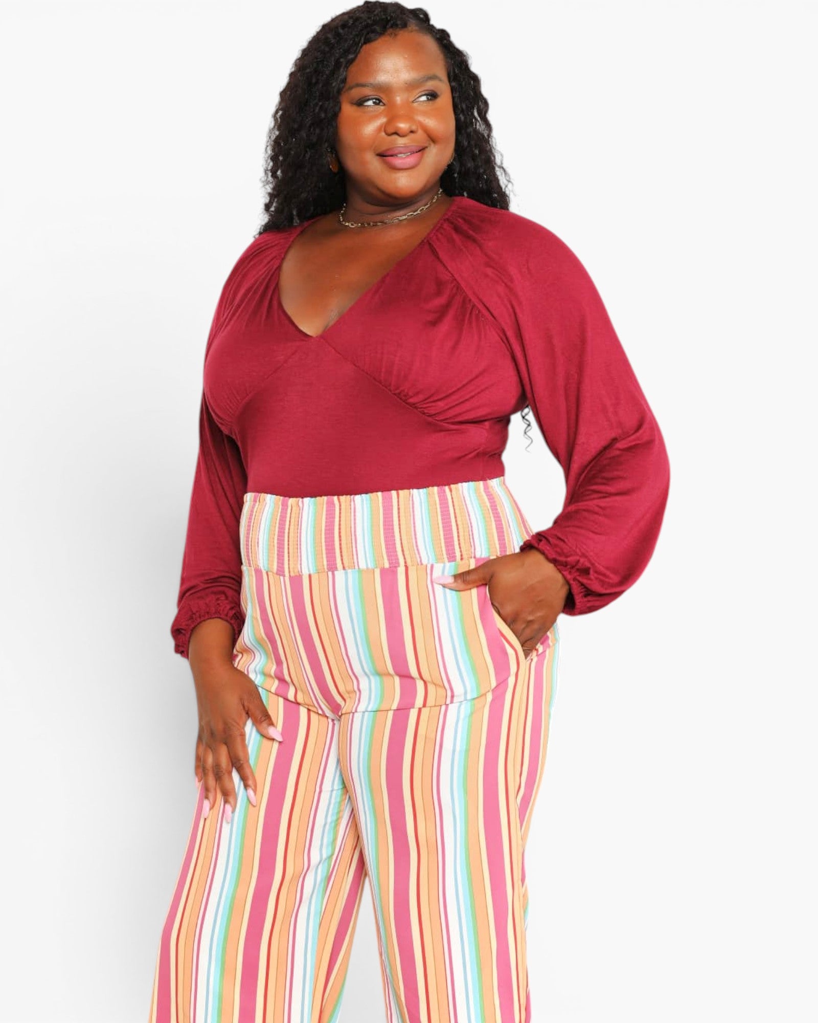 A woman modeling a plus-size outfit featuring a deep burgundy wrap-style top with balloon sleeves and high-waisted, wide-leg trousers with vertical pastel stripes in pink, yellow, blue, and white.