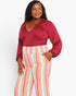 A woman modeling a plus-size outfit featuring a deep burgundy wrap-style top with balloon sleeves and high-waisted, wide-leg trousers with vertical pastel stripes in pink, yellow, blue, and white.