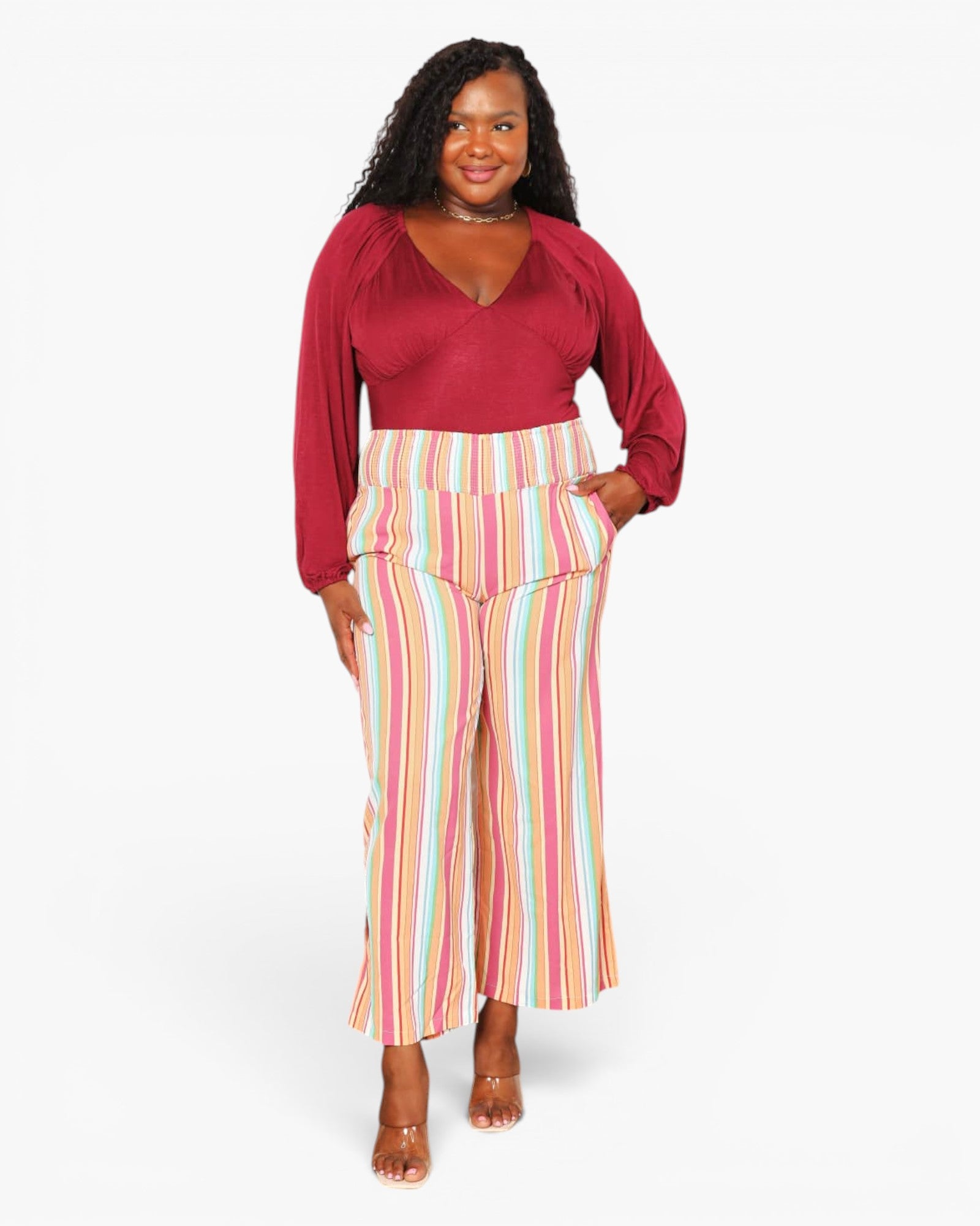 A woman modeling a two-piece outfit consisting of a deep red long-sleeve top and high-waisted, wide-leg cropped pants with vertical pastel stripes in pink, yellow, blue, and white.