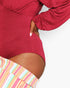 A close-up of a person wearing a long-sleeve deep red bodysuit with a high-cut thigh, paired with pastel striped shorts, against a plain white background.