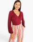 A woman modeling a long-sleeve burgundy top with a gathered V-neckline, paired with colorful vertically striped pants, against a plain light gray background.