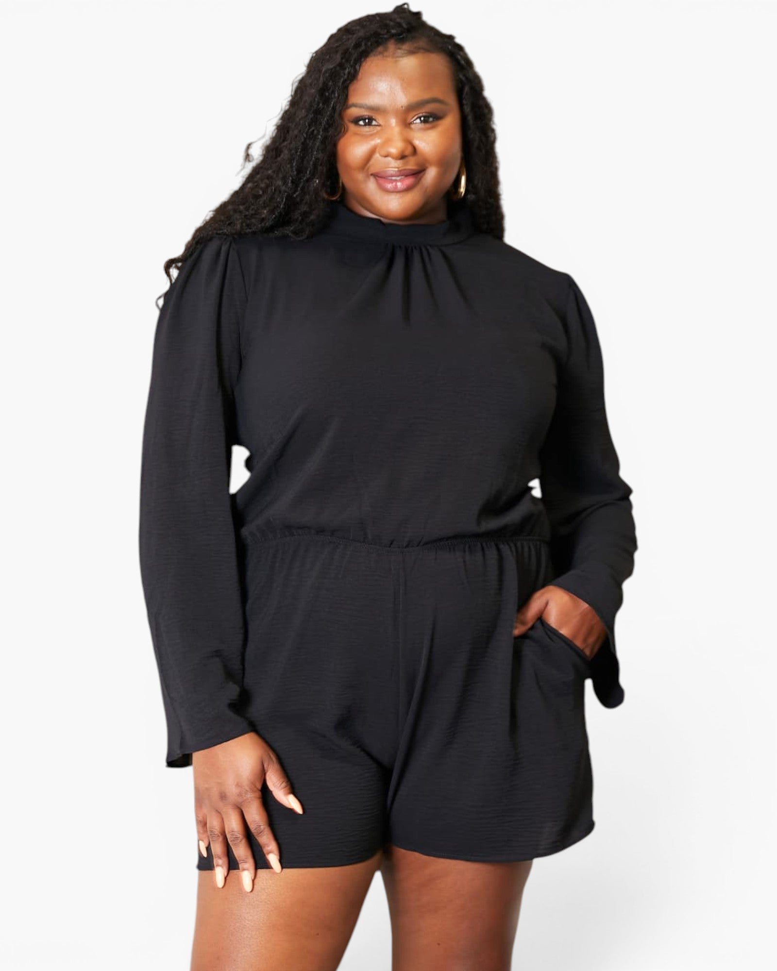 A woman modeling a black long-sleeve romper with a high gathered neckline and side pockets, photographed against a clean white studio background.