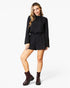 A woman modeling a black long-sleeve romper with a gathered waist, paired with dark brown ankle boots, against a plain white studio background.