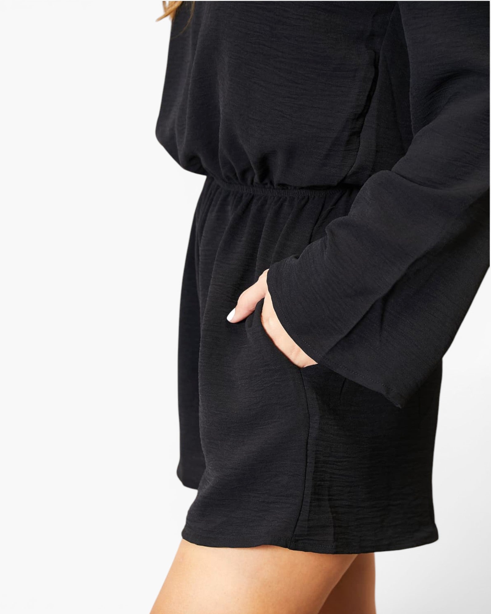 Close-up of a person wearing a black long-sleeve romper or dress with a gathered waist and side pocket, shown against a plain light background.