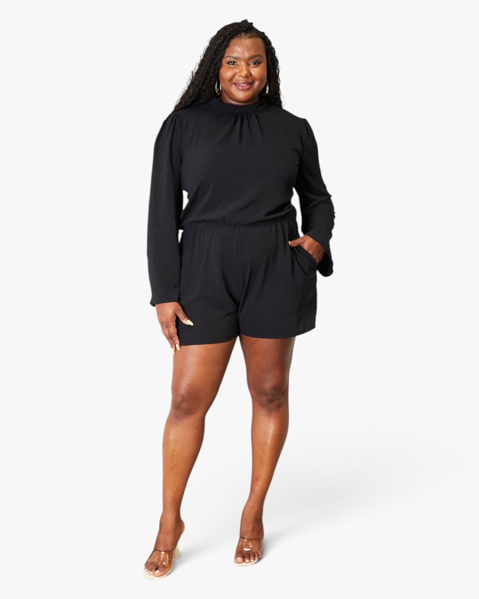 A woman modeling a black long-sleeve romper with a high gathered neckline and side pockets, paired with clear high-heeled sandals, against a plain white studio background.
