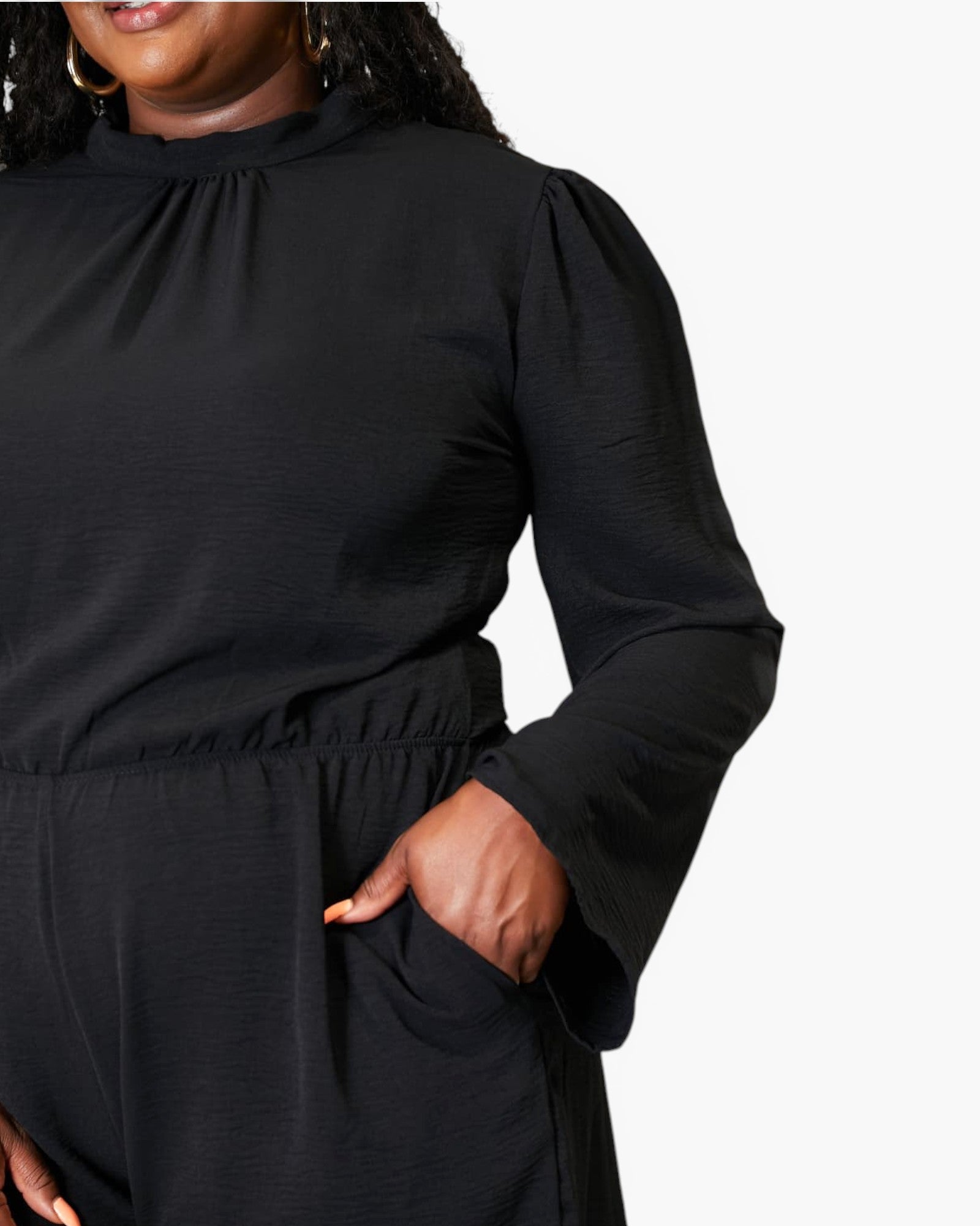 A close-up of a person wearing a black long-sleeve top or jumpsuit with a gathered high neckline, puffed sleeves, and a side pocket, shown against a plain light background.