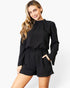 A woman modeling a black long-sleeve romper with a gathered waist and short legs, posed against a clean white studio background.